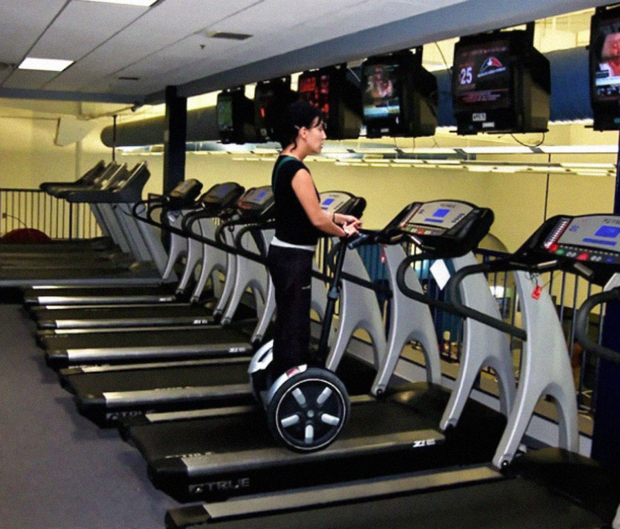 A person rides a Segway on a treadmill in a gym, surrounded by other empty treadmills, with several TVs mounted overhead.