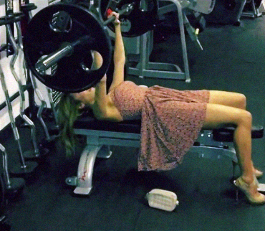 A woman in a pink floral dress lifts a barbell while lying on a gym bench. She is barefoot and her purse is on the floor next to the bench. Gym equipment surrounds her.
