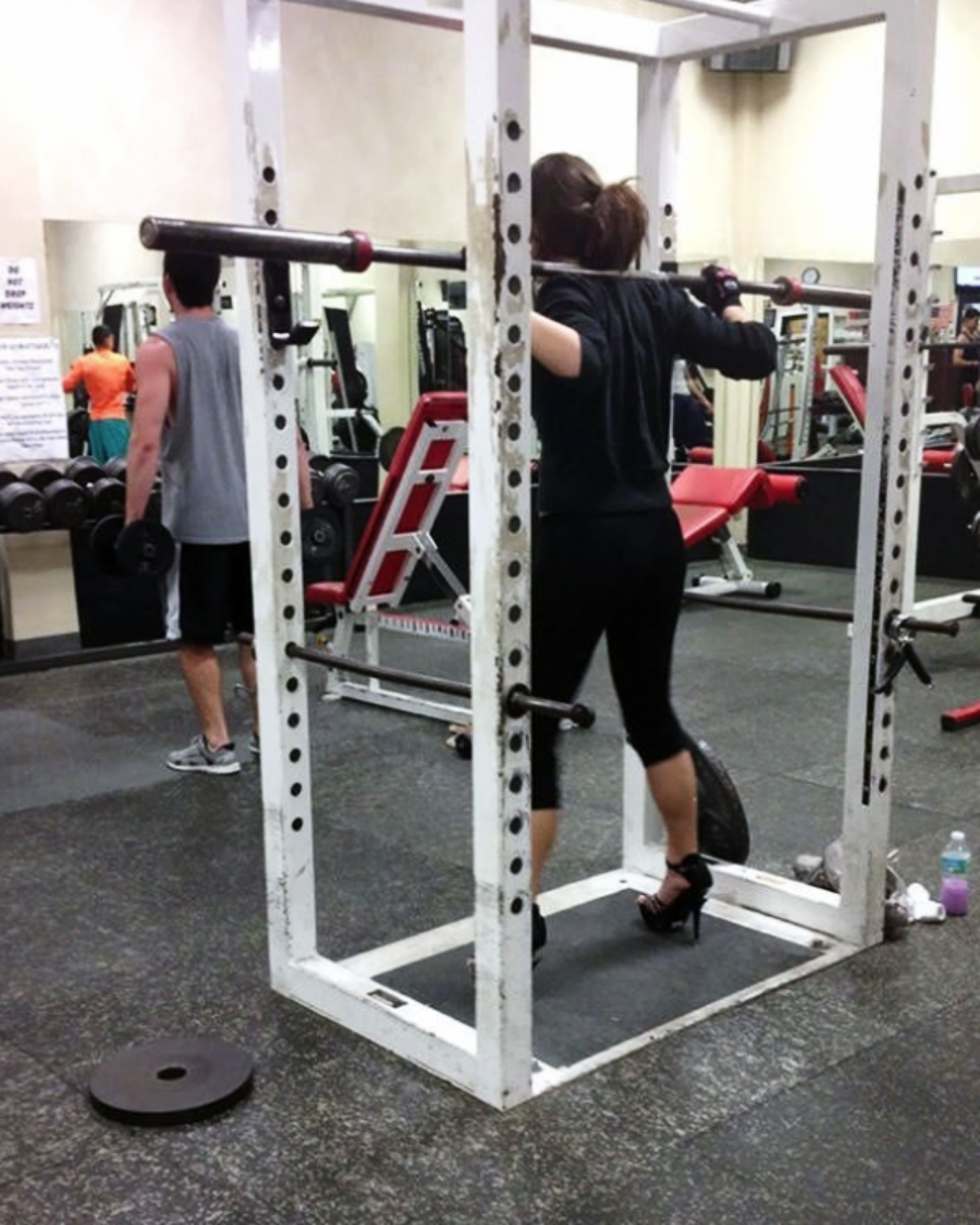 A woman wearing black high heels and workout clothes prepares to lift a barbell in a squat rack at the gym; a man in athletic wear walks nearby.
