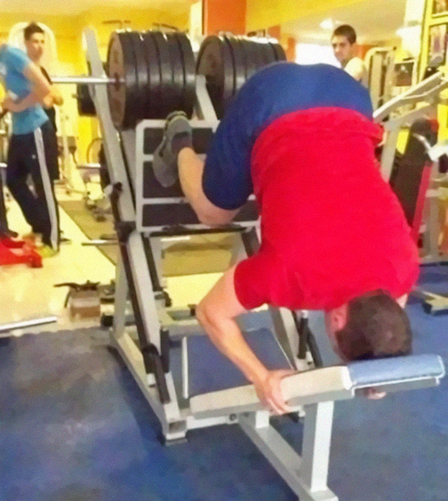 A man wearing a red shirt and blue shorts is using a leg press machine incorrectly, lying face down and pushing the weights upward with his back. Other people in the gym are looking on, some surprised.