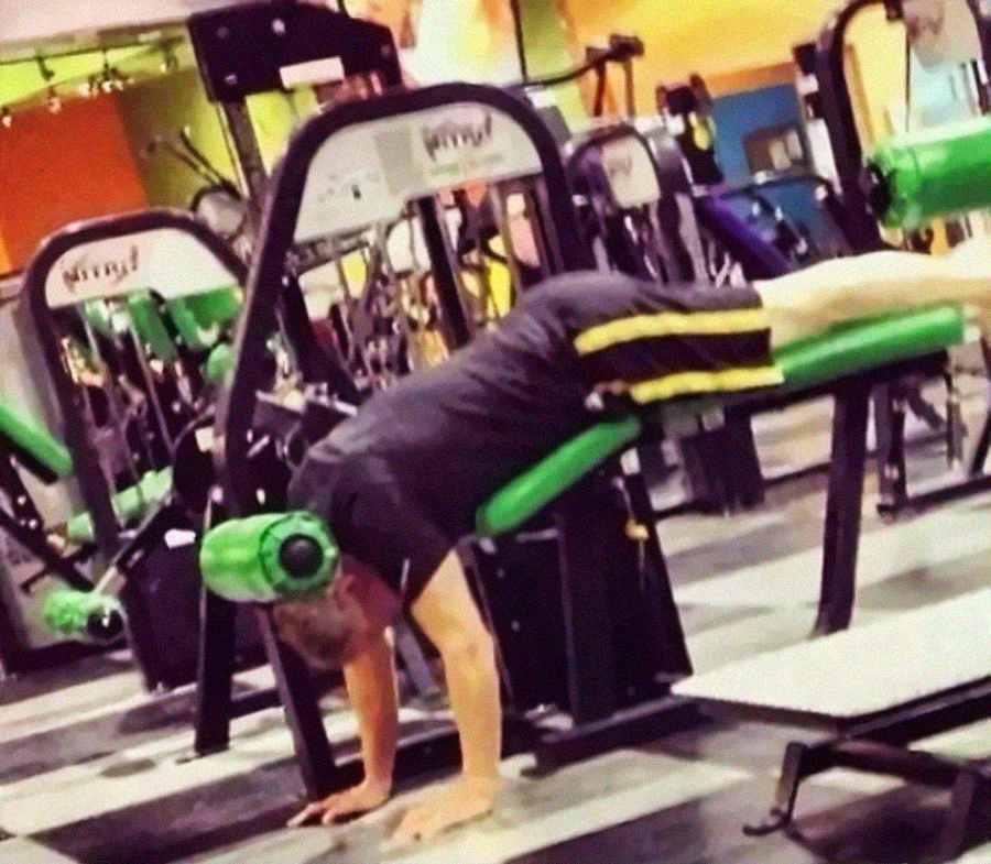A person is upside down on a workout bench in a gym, with their legs on the bench and hands on the floor, appearing to do a handstand or use the equipment incorrectly. Other gym equipment is visible in the background.