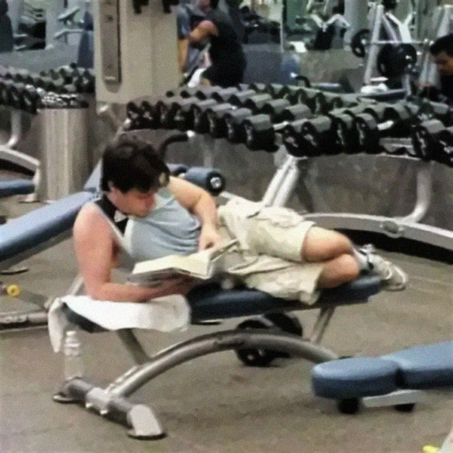 A person is lying on a bench in a gym, reading a book, surrounded by weights and gym equipment. They appear relaxed and are not exercising.