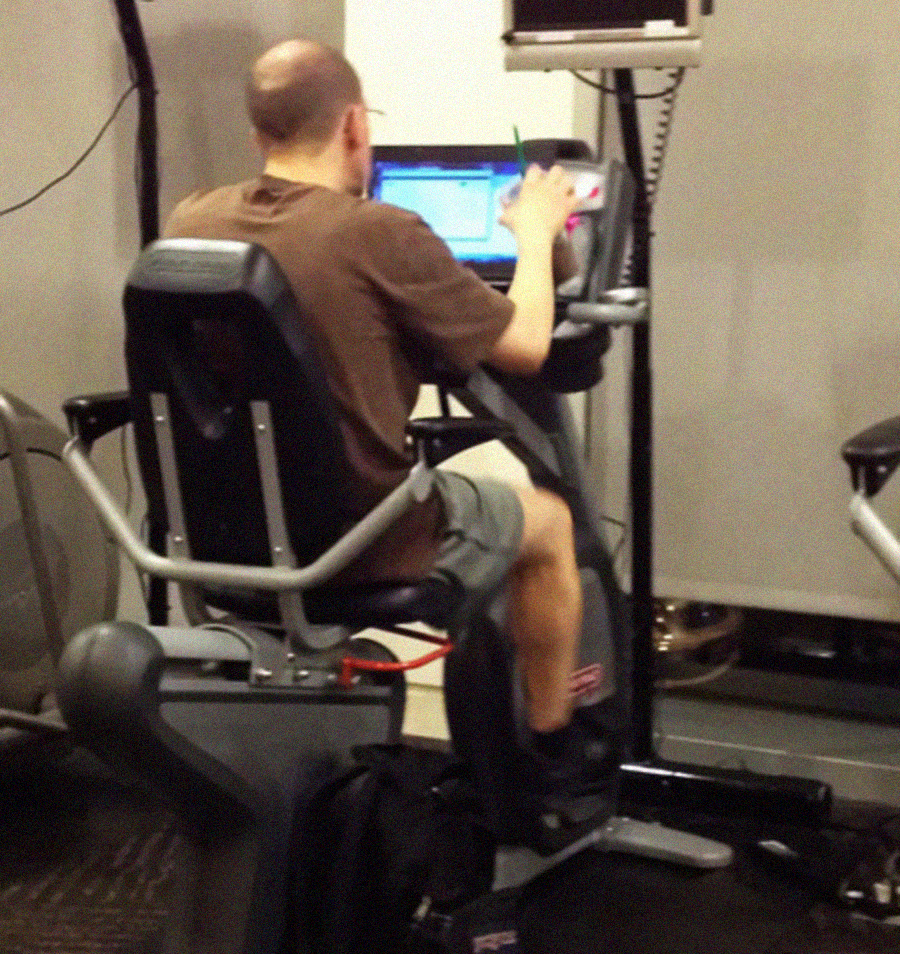 A man sits on a stationary exercise bike in a gym, focusing on a laptop placed on the bike's console while pedaling. He wears a brown t-shirt and gray shorts. The gym equipment and a wall are visible in the background.