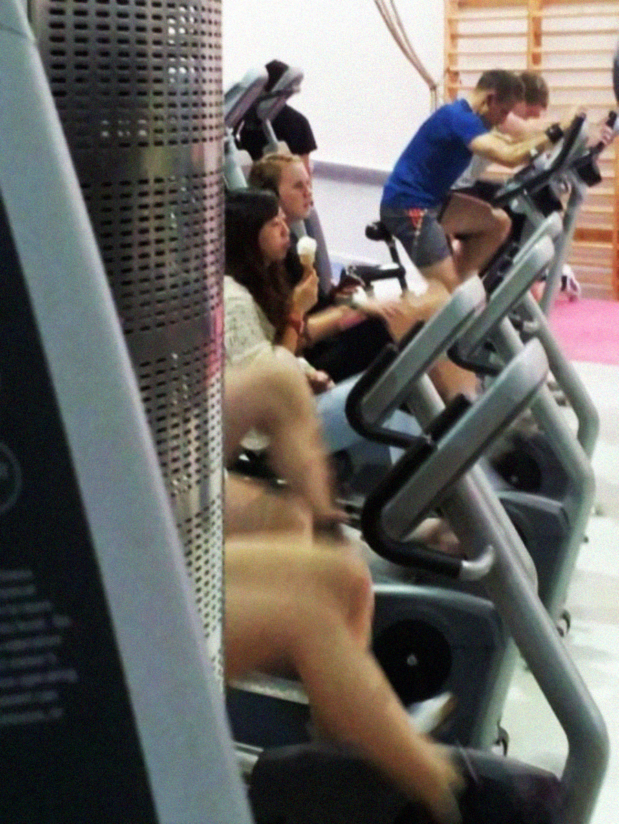 Several people are using exercise bikes at a gym; one person in the middle is casually eating an ice cream cone while pedaling. The atmosphere appears relaxed and informal.