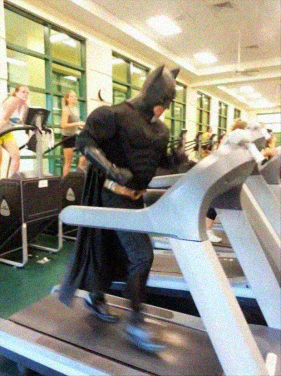 A person dressed in a full Batman costume runs on a treadmill in a gym, while other people exercise on nearby machines in the background.