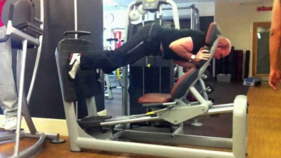 A man at the gym is incorrectly using a leg press machine by lying face down on the seat and pushing the platform with his feet, instead of sitting upright as intended.