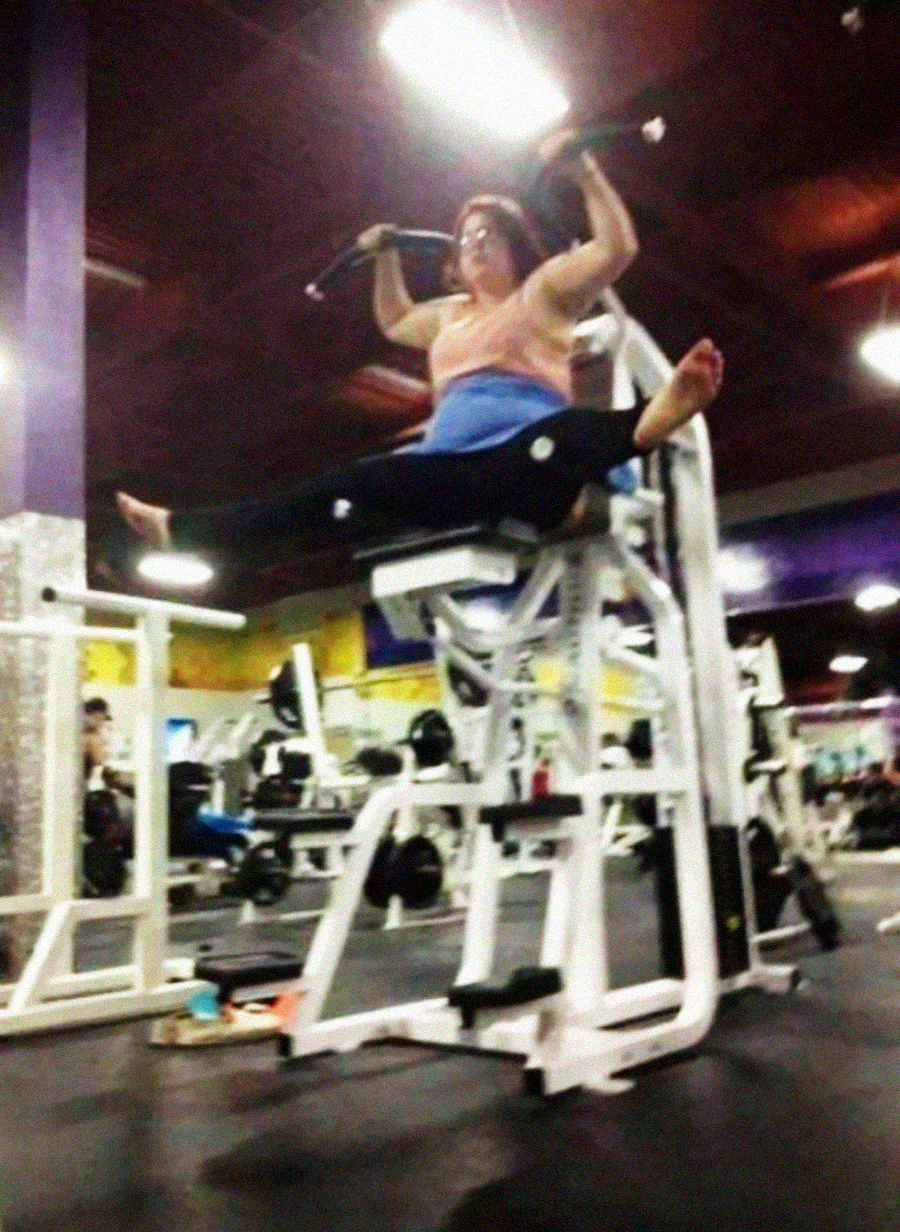 A woman at the gym performs an impressive split while using an exercise machine, raising her legs high in the air and holding onto handles, surrounded by gym equipment.