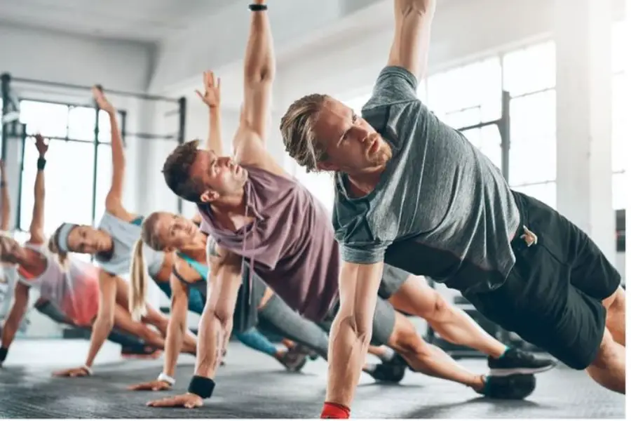 A group of people exercising in a gym, performing side plank poses on the floor, with arms extended upward and bodies aligned, in a bright, spacious room.