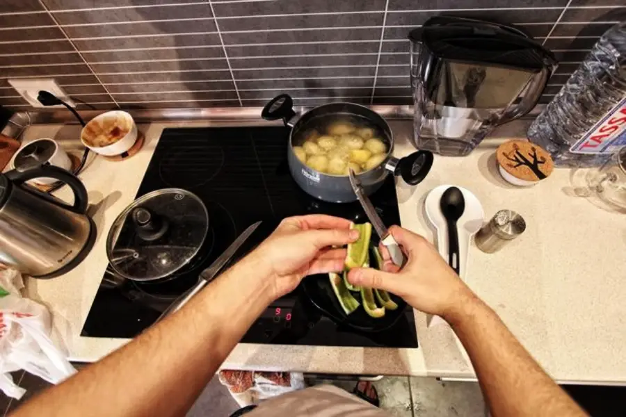 A person peels a cucumber over a stove where a pot of boiling potatoes and a covered pan are cooking. Various kitchen items, including a kettle, water filter, and utensils, are on the countertop.