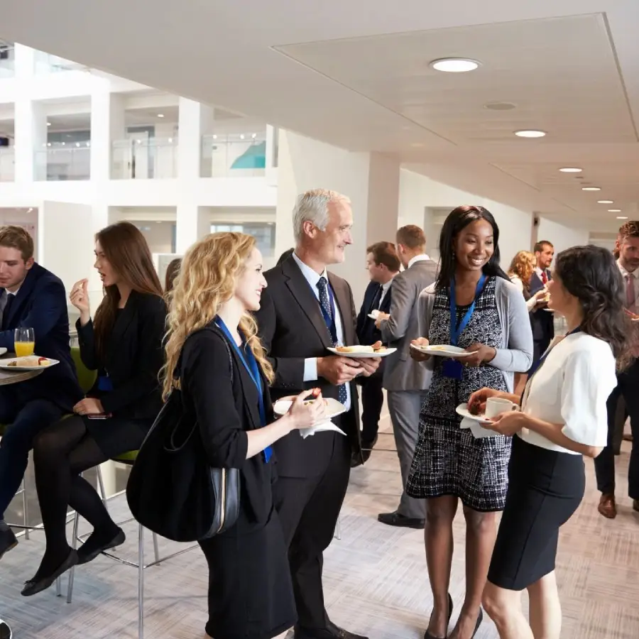 A group of professionals in business attire are standing and chatting while holding plates of food at a modern indoor networking event or conference.