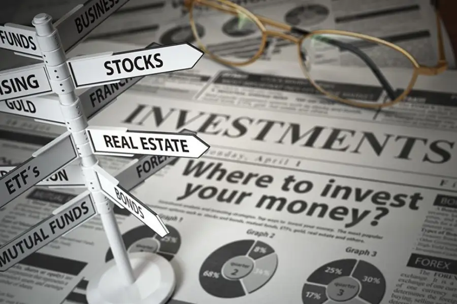 A newspaper with the headline “Where to invest your money?” lies under a signpost showing investment options like stocks, bonds, and real estate. A pair of eyeglasses rests on the newspaper.