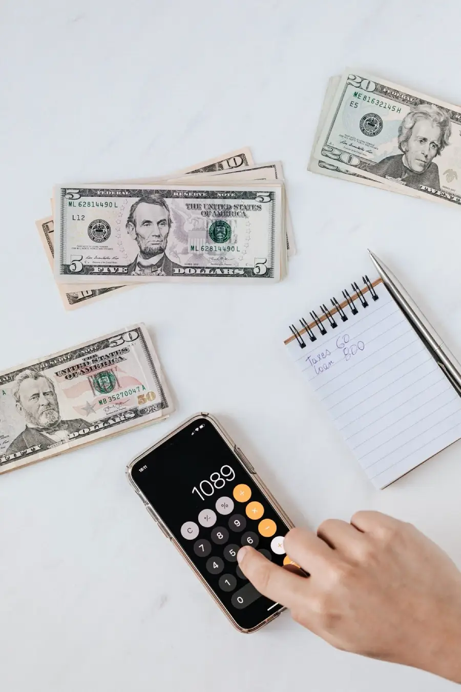 A hand using a calculator app on a smartphone, with stacks of US dollar bills and a notepad with financial notes on a white surface.
