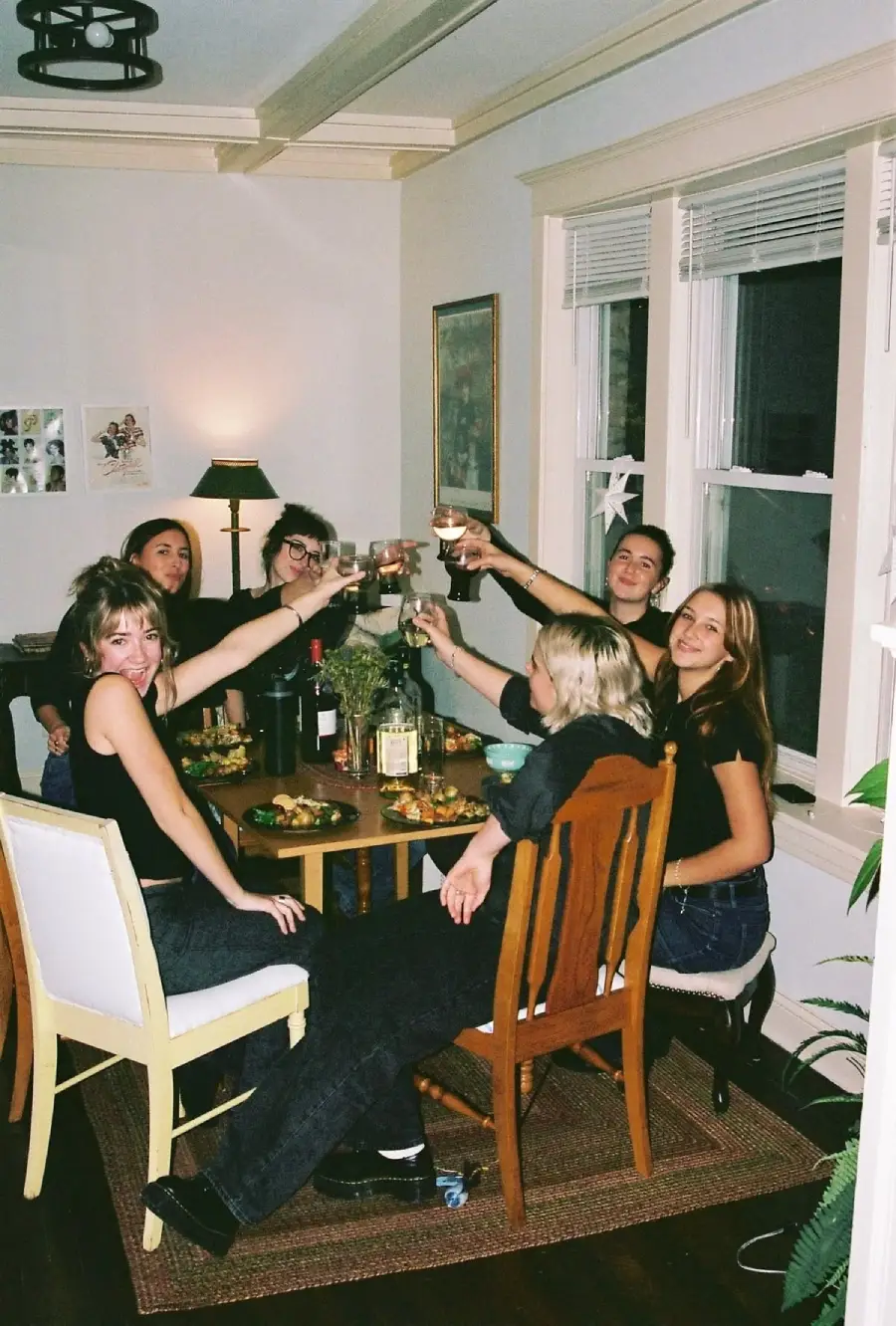 Six people sit around a wooden dining table, raising glasses in a toast and smiling. The table is set with food, drinks, and bottles. The room has white walls, large windows, and framed art on the wall.