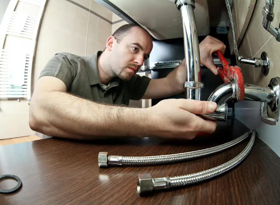 A man is fixing plumbing under a sink using a red pipe wrench. Flexible metal hoses and a black washer lie on the wooden countertop nearby. Sunlight comes through blinds in the background.