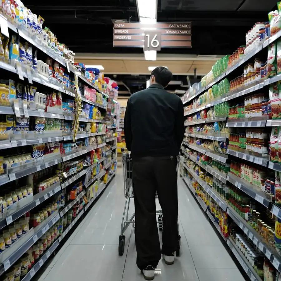 A person pushes a shopping cart down a supermarket aisle lined with various food products on both sides, under a sign labeled "16" for pasta, spreads, preserves, sauce, and tomato sauce.