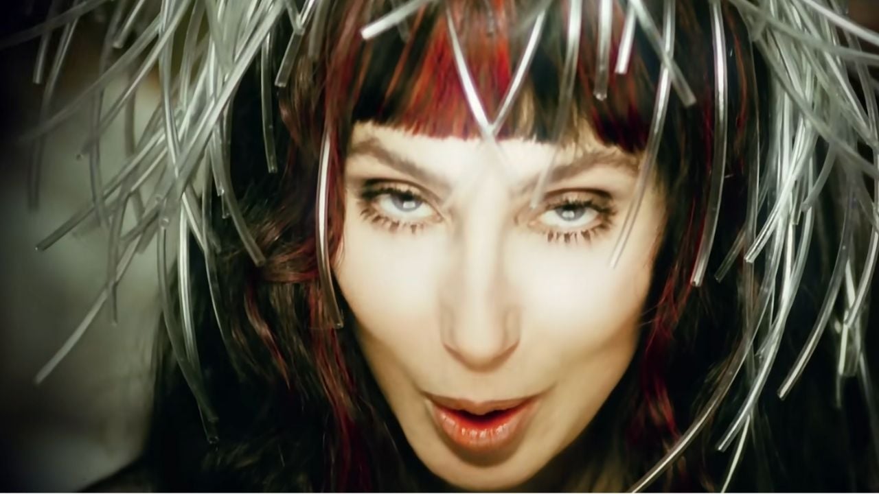 A person with dramatic eye makeup and red streaks in dark hair wears a spiky, metallic headpiece, looking directly at the camera with a bold expression.