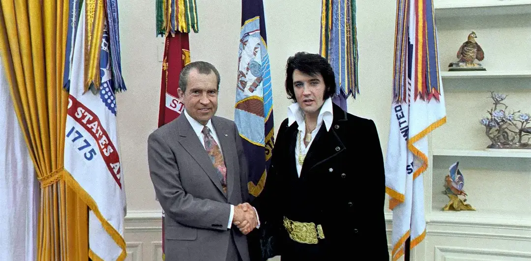 Two men stand in an office, shaking hands. One wears a suit and tie, the other a black outfit with a large belt. Colorful flags and yellow curtains are in the background, along with shelves holding decorative items.