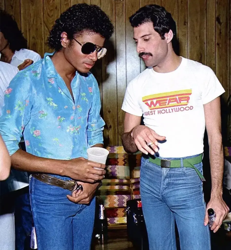 Two men stand together talking. One wears sunglasses, a blue floral shirt, and jeans, holding a cup. The other wears a white t-shirt with "WEST HOLLYWOOD," blue jeans, and a green belt, gesturing with his hand.