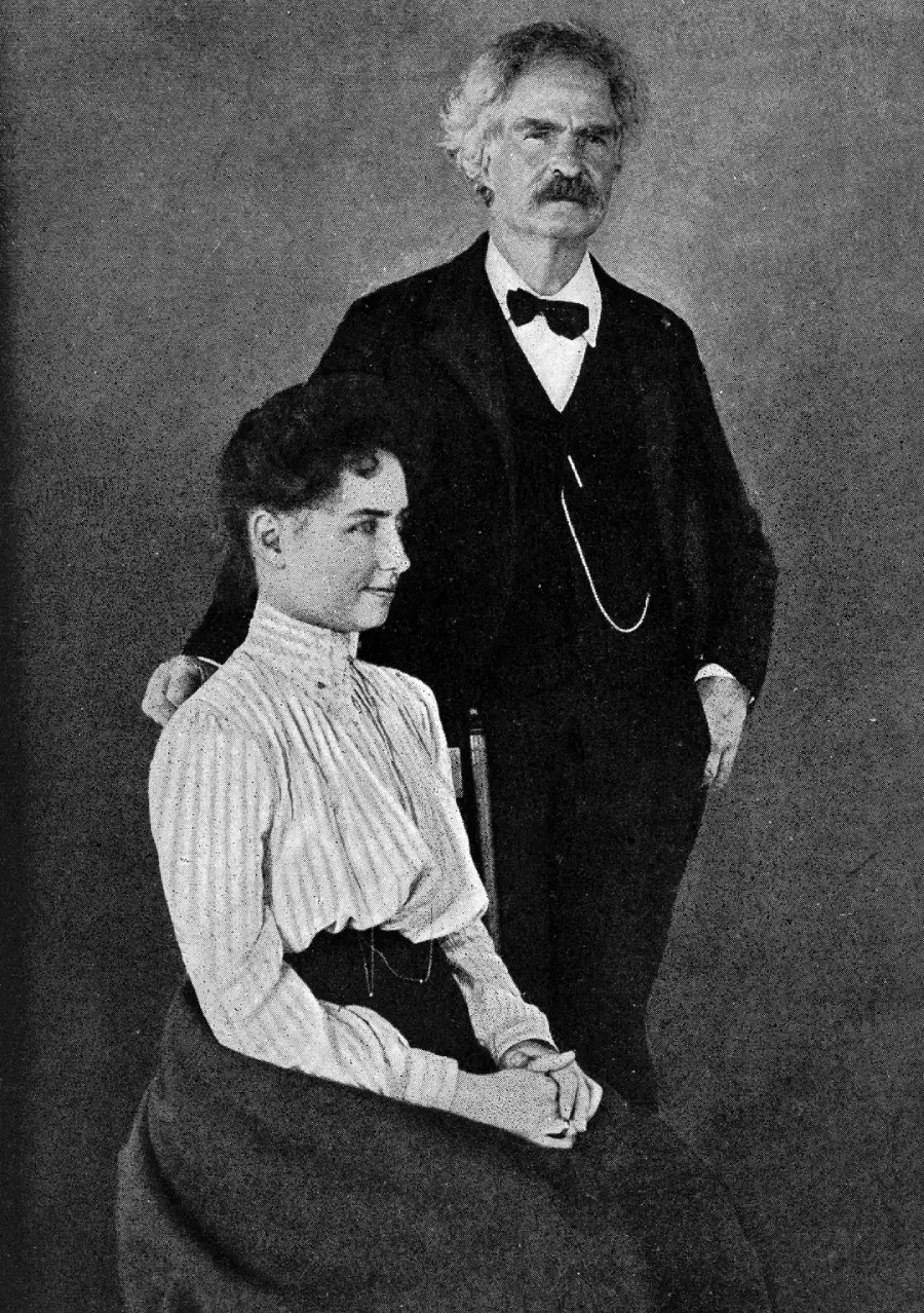 A black-and-white photo of a seated woman in a high-collared blouse and long skirt, with a mustachioed man standing behind her in a suit, bow tie, and glasses, resting one hand on her chair.