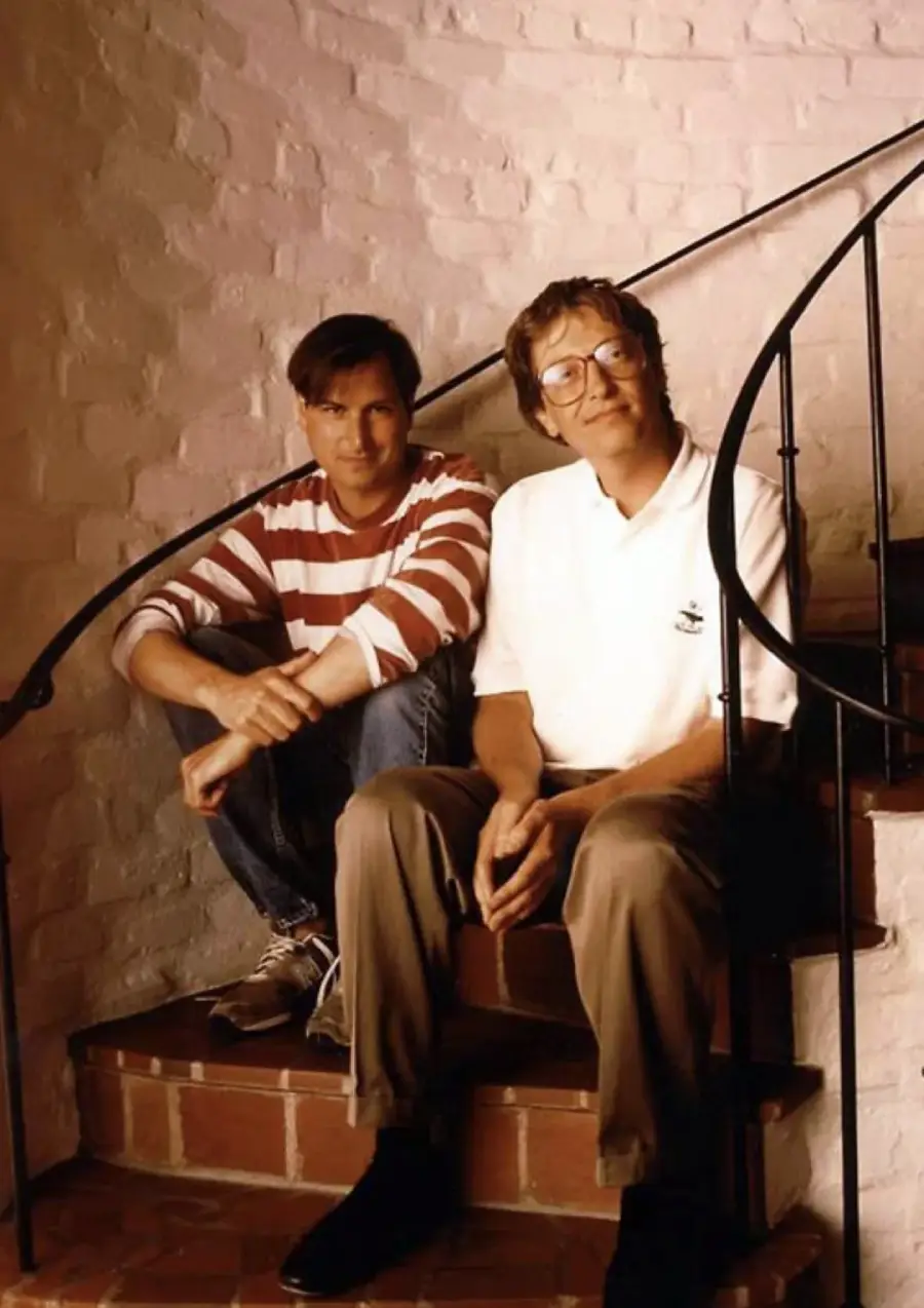 Two men sit on a brick staircase against a white brick wall. One wears a striped shirt and jeans, while the other wears glasses, a white polo shirt, and khaki pants. Both appear relaxed and are looking at the camera.