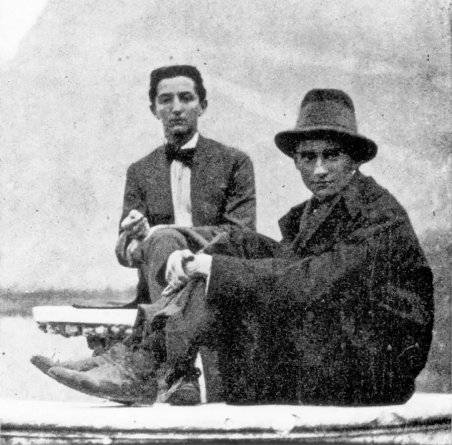 Two young men in vintage clothing pose outdoors. One sits cross-legged in a hat and coat, facing the camera, while the other sits behind him, dressed in a bow tie and jacket, with a blurred background. The image is in black and white.