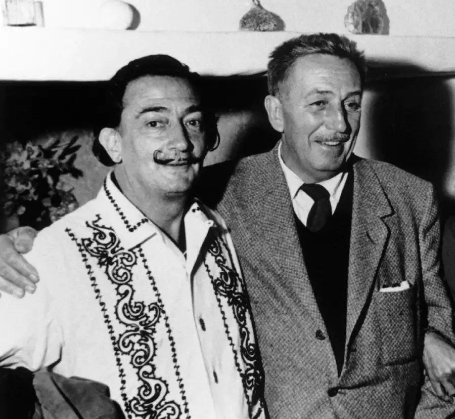 Two smiling men with mustaches stand side by side, one wearing an embroidered shirt and the other in a suit jacket and tie, with arms around each other's shoulders in a black-and-white photo.