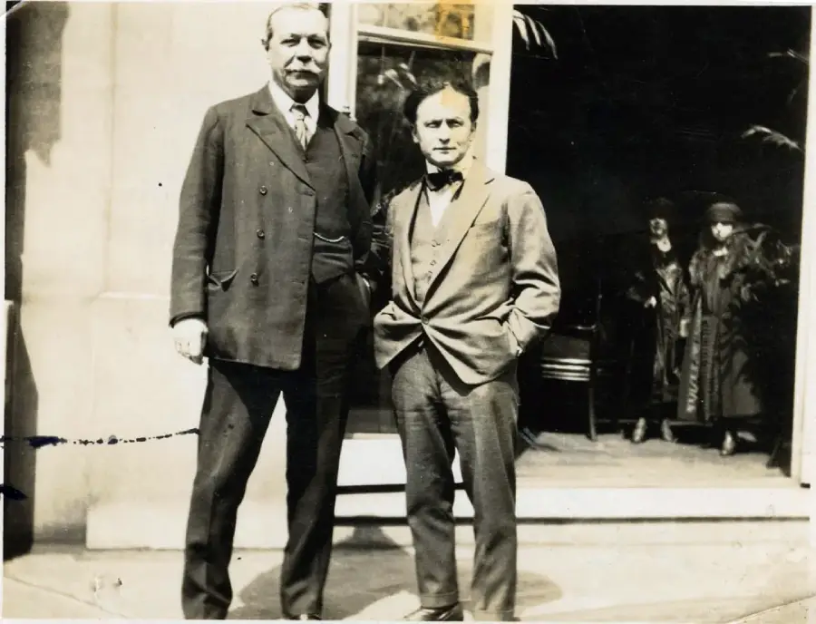 Two men in suits stand outside a building, one much taller than the other. In the background, two women in coats and hats are visible near the entrance. The photo appears to be old and in black and white.
