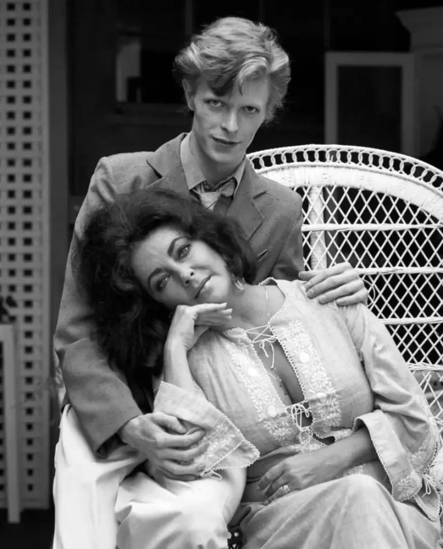 A young man in a suit sits on a wicker chair, gently resting his hands on the shoulders of a woman in a light, laced dress, who leans her head against him and gazes thoughtfully forward. The image is in black and white.