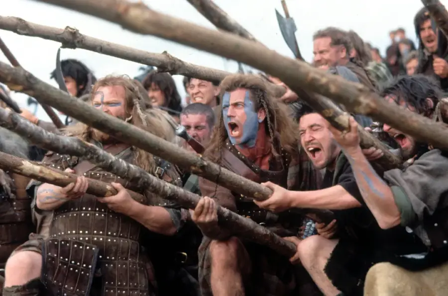 A group of men in medieval battle attire with blue face paint shout fiercely, holding long wooden spears pointed forward, appearing ready for battle on a grassy battlefield.