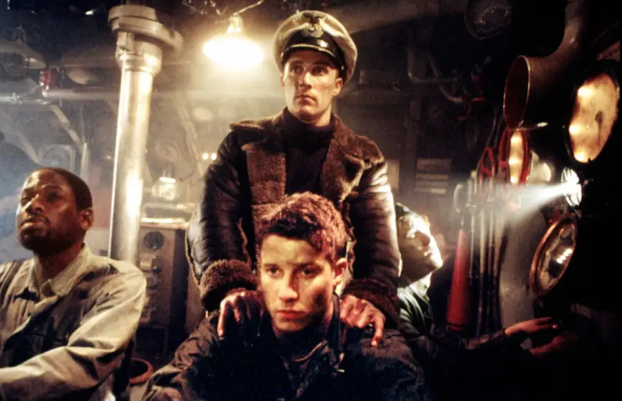 Four men in a dimly lit submarine control room, one wearing a captain's hat and coat, stands behind another with his hands on his shoulders. The others watch tensely, surrounded by pipes and machinery.