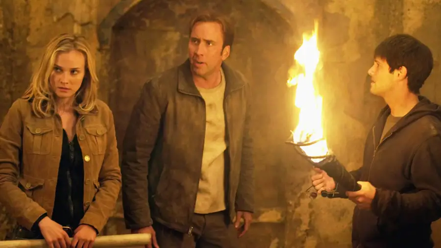 Three people stand in a dimly lit, ancient stone room. One holds a flaming torch. The man in the center looks startled, while the woman and the other man appear tense and alert.