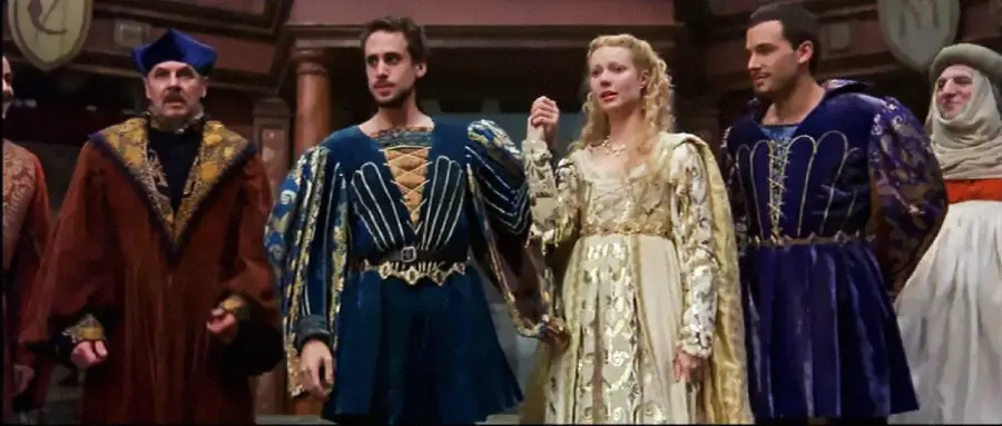 A group of people in elaborate Renaissance-style costumes stand together on a stage, with a woman in a gold gown holding hands with a man in a blue tunic; others wear richly decorated robes and hats.
