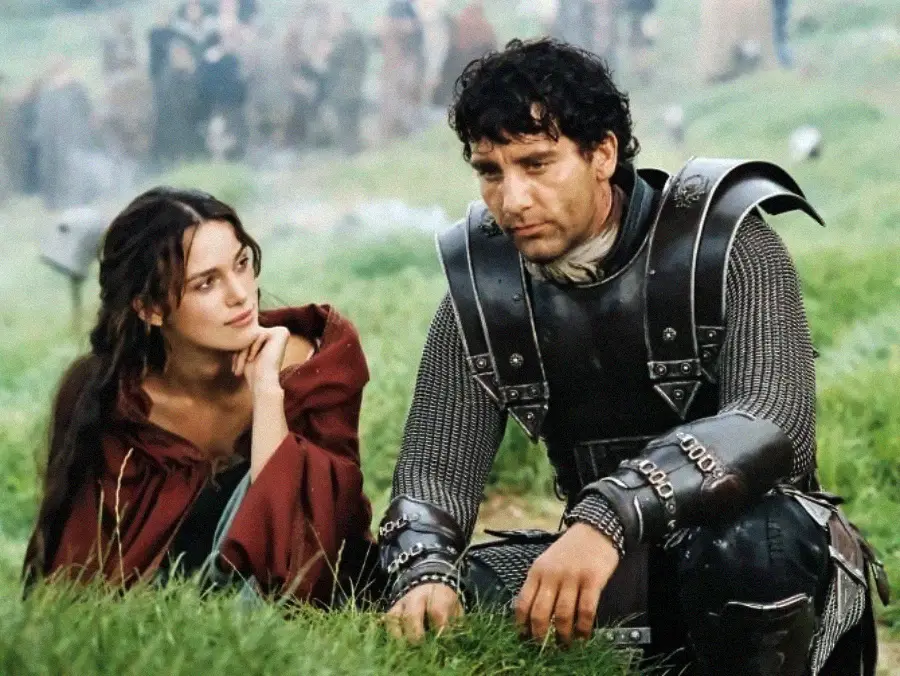 A woman in a red cloak sits in grass, gazing thoughtfully at a man in medieval armor who is sitting beside her. Both appear serious, with a blurred group of people in the background.