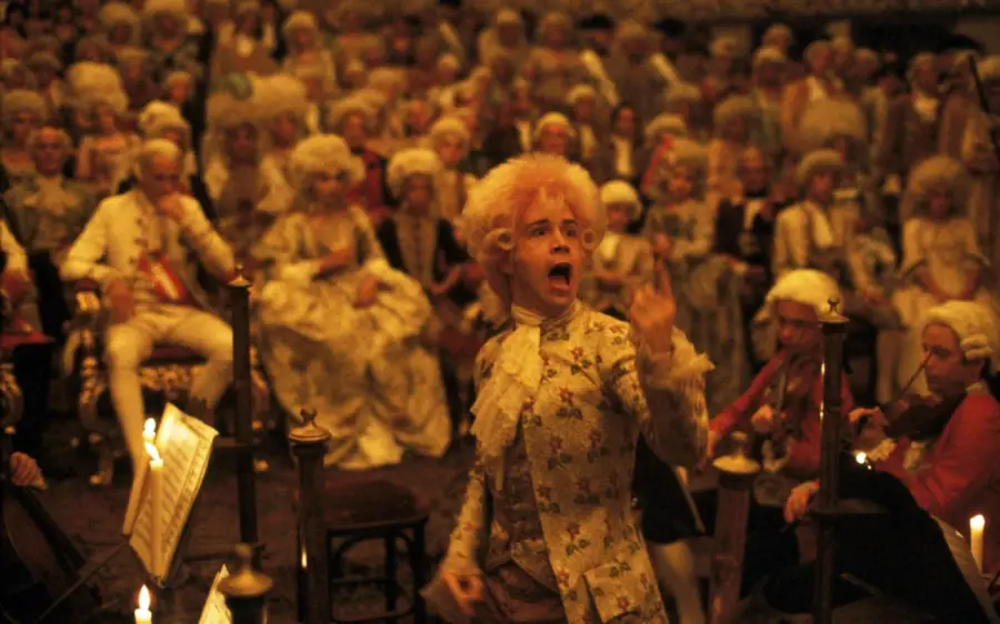 A man in 18th-century attire passionately conducts an orchestra in a candlelit room, surrounded by an audience in elaborate wigs and period costumes. The scene is warm-toned and crowded with figures watching intently.