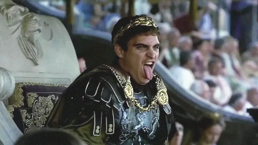 A man in ornate Roman armor and a gold laurel crown sits on a throne and sticks out his tongue, appearing to mock or taunt, with a blurred crowd in the background.