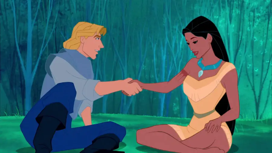 A blonde man and a woman with long black hair sit on grass in a forest, smiling and shaking hands. The woman wears a tan dress and turquoise necklace; the man wears a light blue shirt and dark pants.