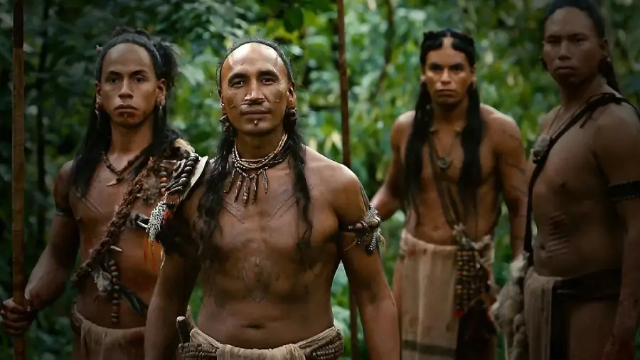 Four Indigenous men with long hair and tribal jewelry stand in a lush, green forest. They are bare-chested and hold spears, looking intently ahead with serious expressions.