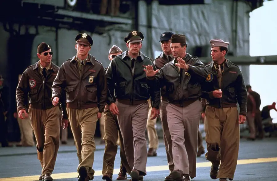 A group of men in military uniforms walk together on an aircraft carrier deck, some wearing bomber jackets and hats, engaged in conversation against a blurred industrial background.