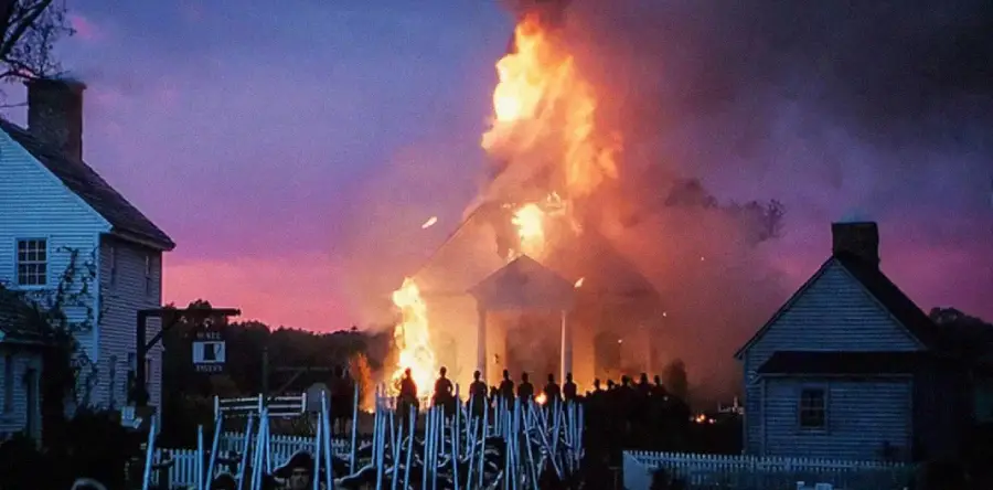 A large fire engulfs a white building at dusk, with flames and smoke rising into the sky. Silhouetted people stand in front, watching the blaze, while nearby houses and a picket fence are visible.