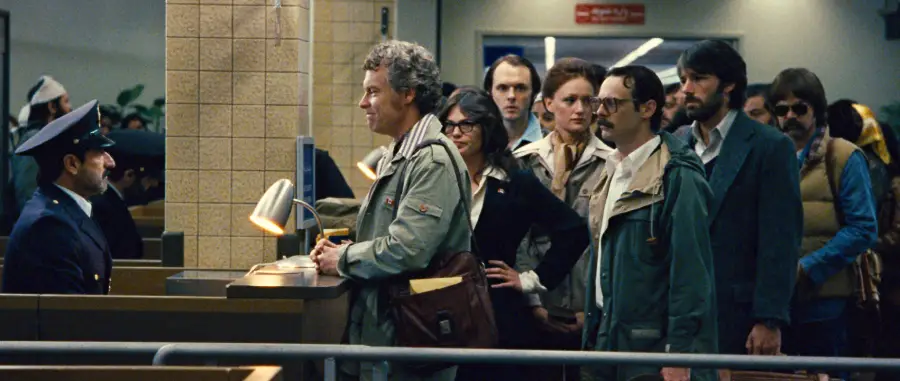 A group of people stand in line at an airport counter, with one man speaking to a uniformed officer while others wait behind him. The setting appears tense and serious.