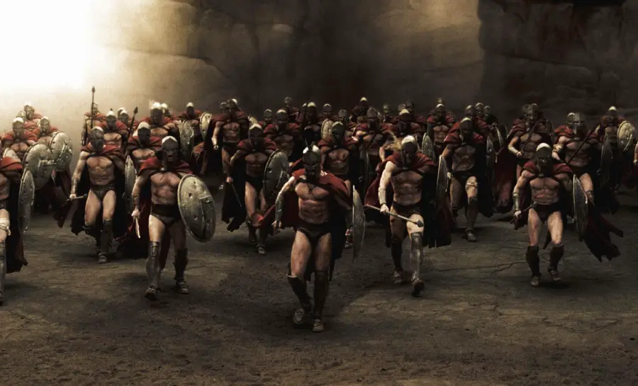 A group of Spartan warriors wearing red capes, helmets, and armor march forward with shields and spears in a dramatic, rocky setting. The scene appears intense and cinematic.