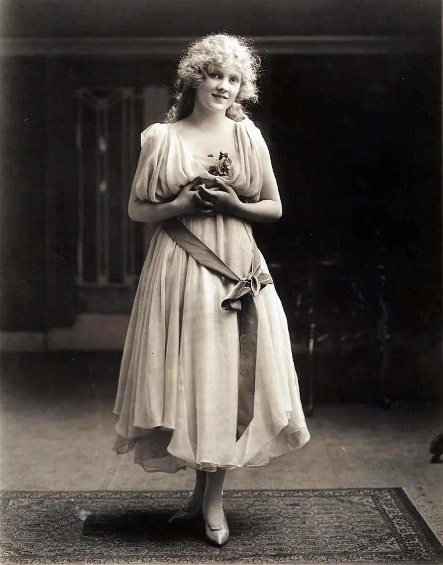 A young woman with curly hair stands on a rug, wearing a light, flowing dress with a sash tied at her waist. She holds a small object in her hands and smiles softly, posing for a vintage, black-and-white photograph.