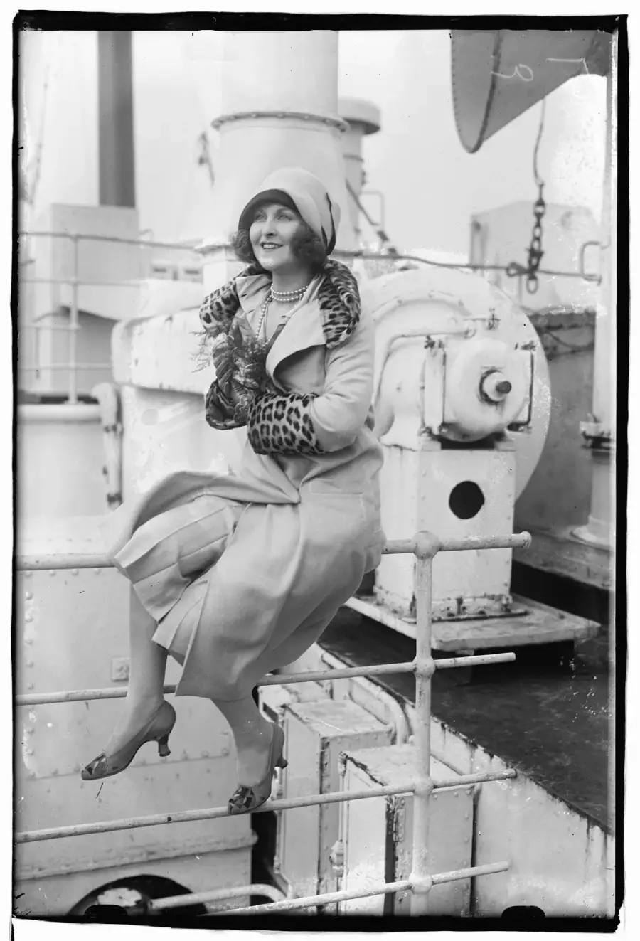 A woman in a cloche hat and elegant dress with a leopard-print collar sits on a ship railing, smiling and holding a lobster. Industrial ship equipment is visible in the background. Black and white photo.