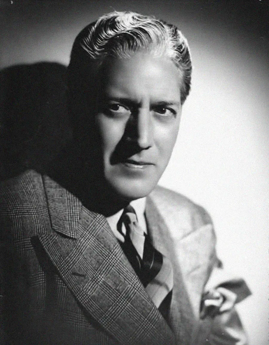 Black and white portrait of a man wearing a suit and tie. He has wavy, slicked-back hair and a serious expression, looking slightly to the side. The lighting creates a dramatic shadow behind him.