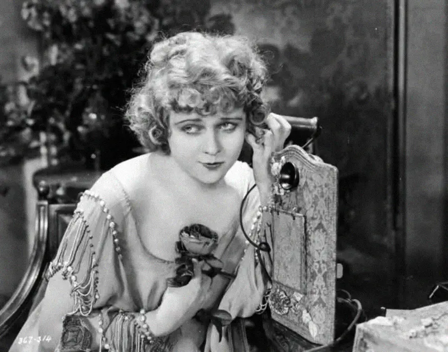 A young woman with curly hair and a vintage dress holds a flower and listens intently to an old-fashioned wall telephone, evoking a scene from the early 20th century.