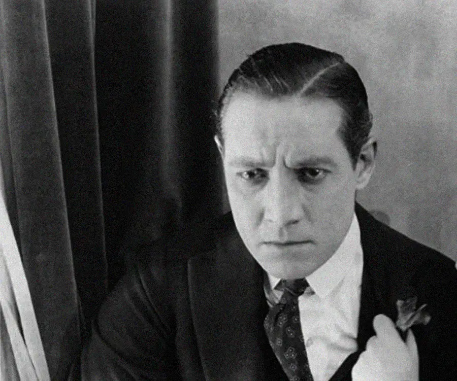 A man in a suit with slicked-back hair sits in front of a curtain, looking down with a serious or concerned expression.