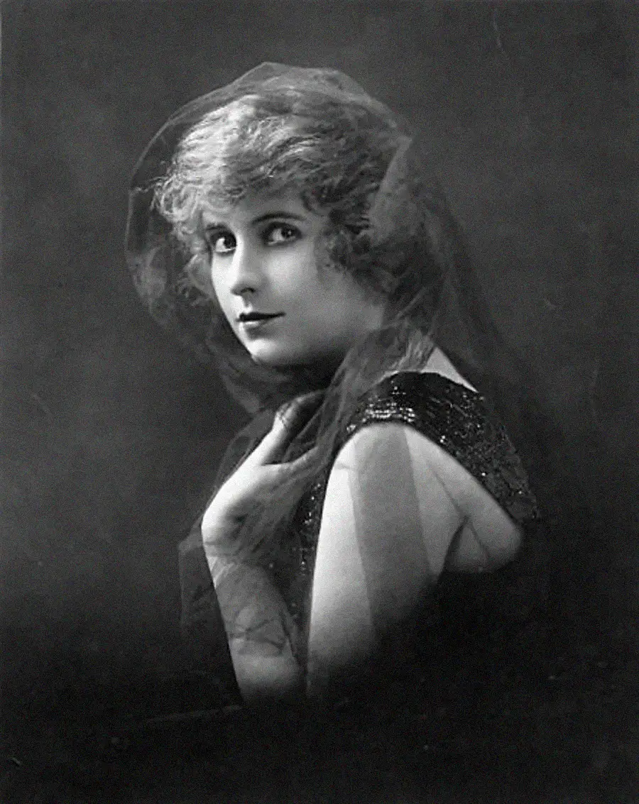 A woman with light curly hair poses in a vintage black-and-white portrait, wearing a dark dress and a sheer veil over her head and shoulders, looking slightly to the side with a soft, elegant expression.