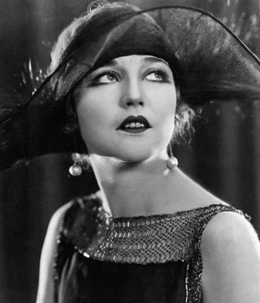 Black and white portrait of a woman wearing a dramatic wide-brimmed hat with a feather, bold lipstick, pearl drop earrings, and a dress with decorative trim, gazing upward with an elegant expression.