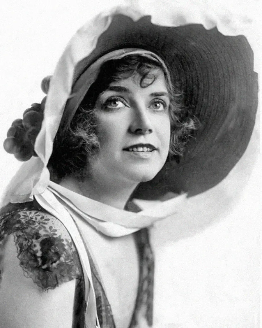 A woman with curly hair gazes upward, wearing a large, wide-brimmed hat adorned with a scarf and lace-accented dress, in a black-and-white vintage portrait.