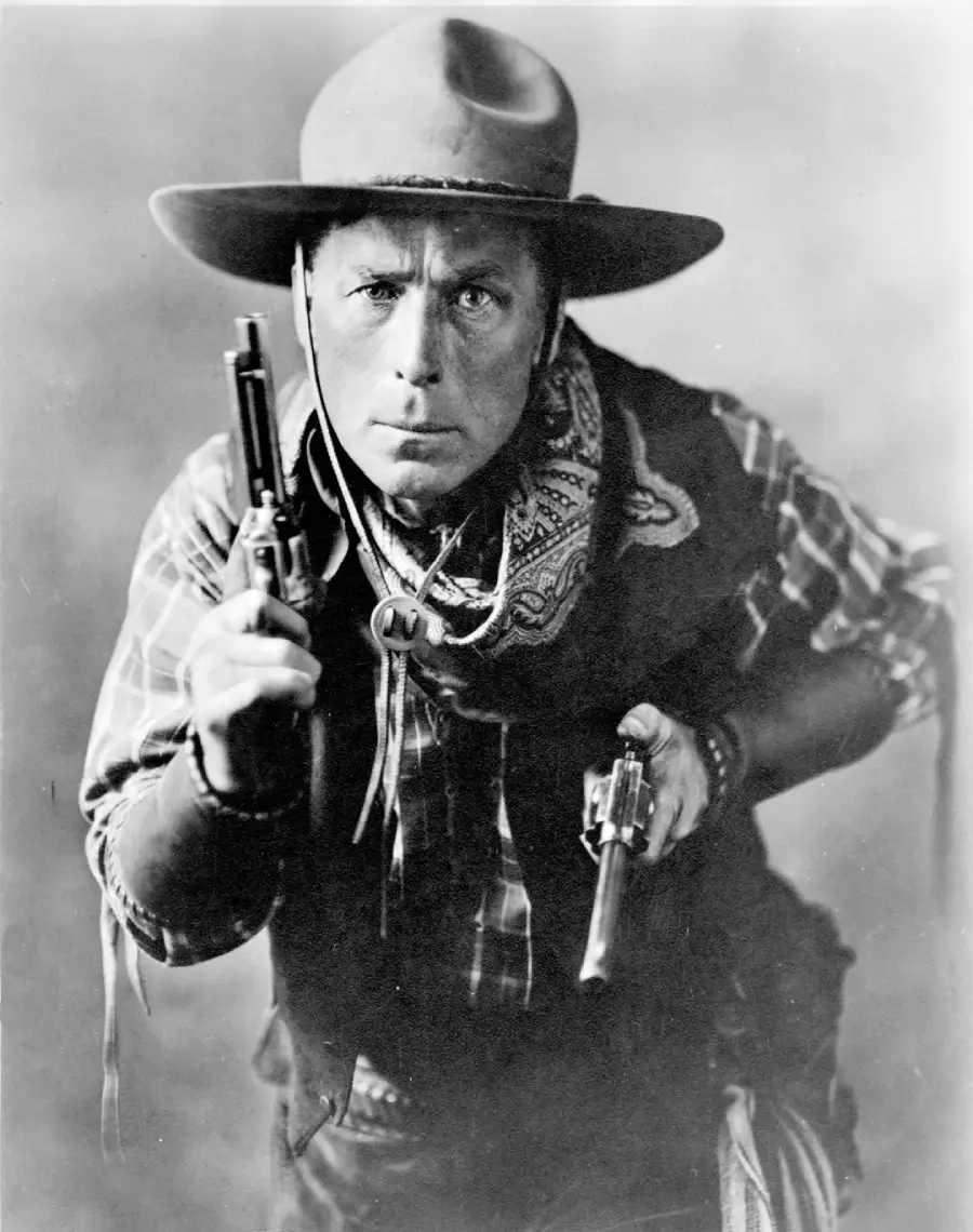 A man dressed as a cowboy in a hat, scarf, and plaid shirt points two pistols toward the camera with a serious expression. The photo is black and white and has an old-fashioned, vintage style.